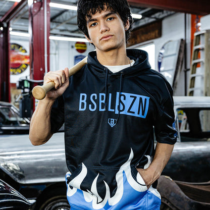Young man holding wooden bat over shoulder in vintage garage, wearing black BSBLSZN short sleeve hoodie with blue flame hem and blue flame mesh shorts.