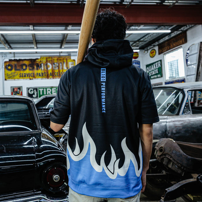 Back view of black BSBLSZN short sleeve hoodie showing vertical BL101 Performance text and blue and white flame hem, model holding bat in vintage garage.