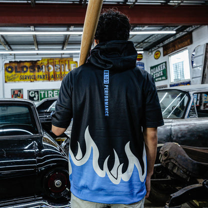 Back view of black BSBLSZN short sleeve hoodie showing vertical BL101 Performance text and blue and white flame hem, model holding bat in vintage garage.