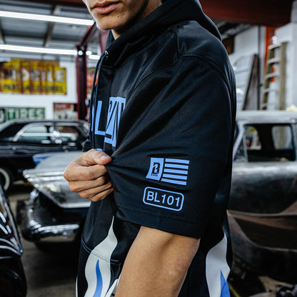 Side close-up of blue BL101 flag and BL101 sleeve hits on black short sleeve hoodie, vintage garage in background.