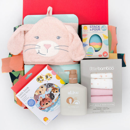 A baby hamper gift set including a pink bunny hooded towel, bath toys, a bath book, and washcloths, all displayed in a red gift box. Price: 135