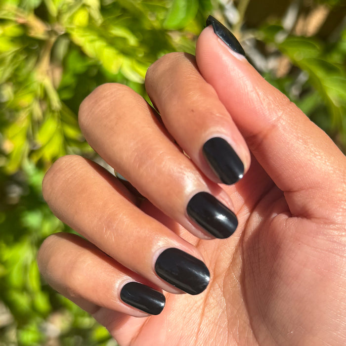 Cateye | Extra Short | Squoval - Tab Press-On nails in glossy deep black shown on hand outdoors with green foliage background