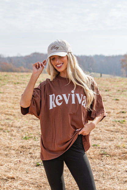 Woman wearing a brown 'Revival' shirt and beige cap in an open field.