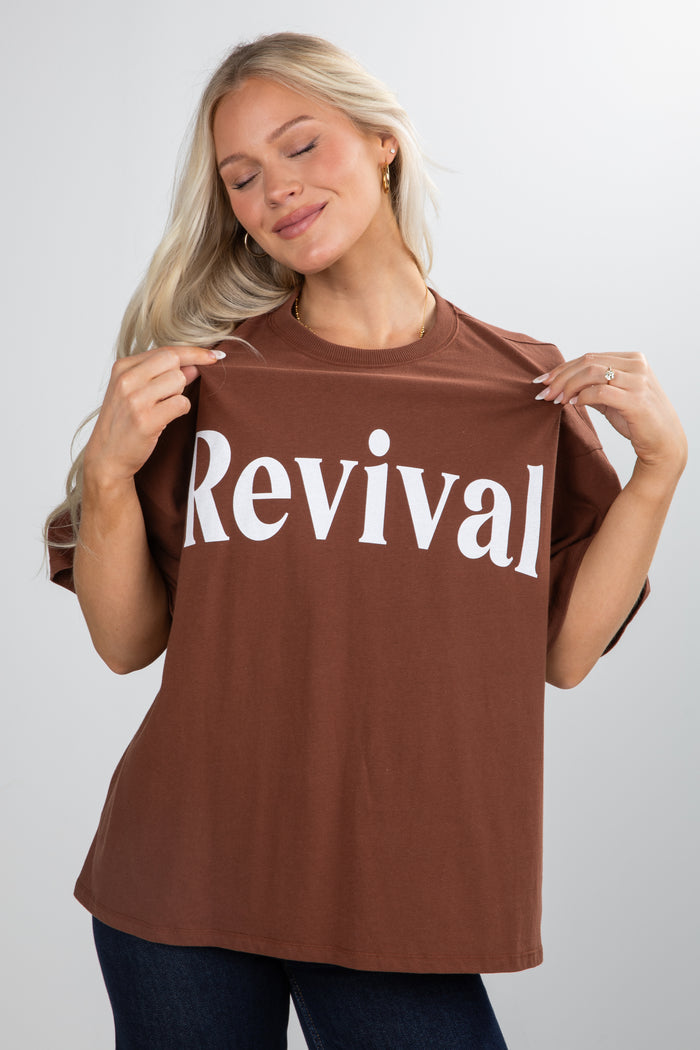 Woman wearing a brown t-shirt with 'Revival' printed on it against a plain background