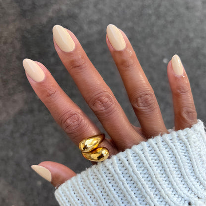 Hand with almond-shaped nails painted in creamy neutral tan Coffee Milk polish wearing gold ring and white knit sweater