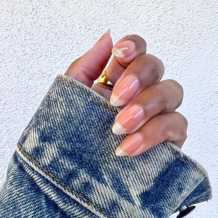 Hand with natural nails polished in Crystal clear coat holding a denim jacket sleeve showing a glossy manicure