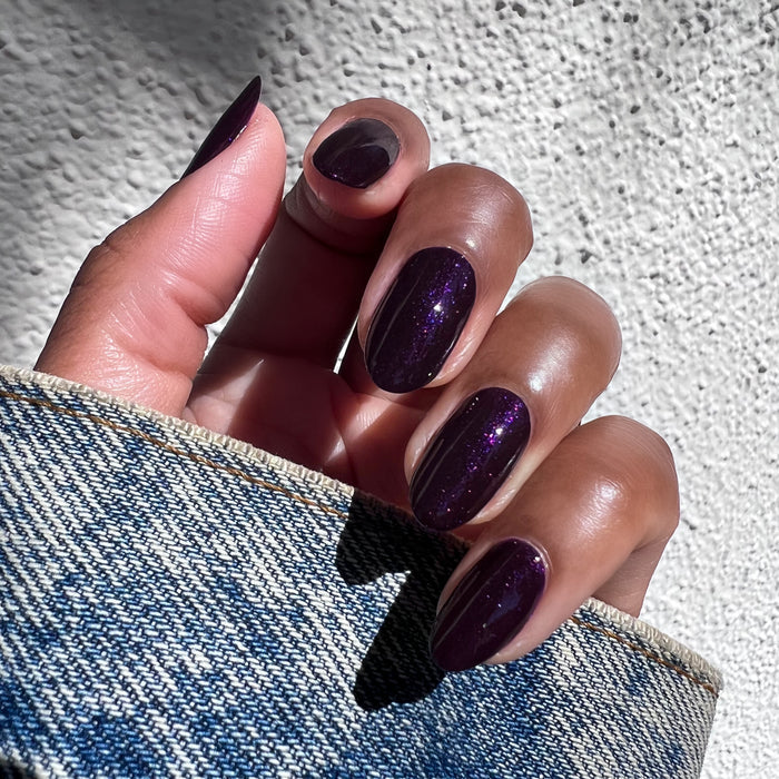 Hand with nails painted in shimmering dark violet Enchanted polish shown against denim fabric for a stylish mani