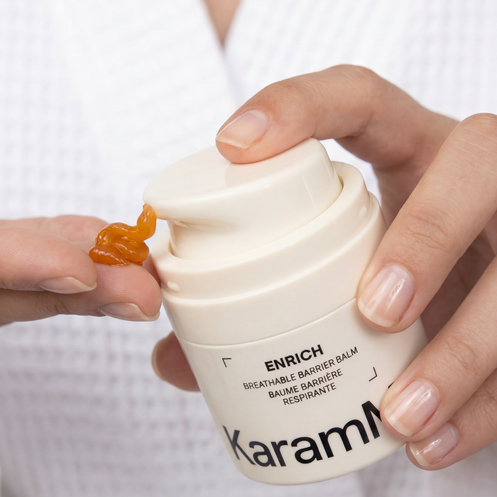 A close-up of a person's hands holding a white pump bottle of Karama Enrich Breathable Barrier Balm, with a small dollop of amber-colored balm shown on the tip of the finger against a white waffle-textured background.