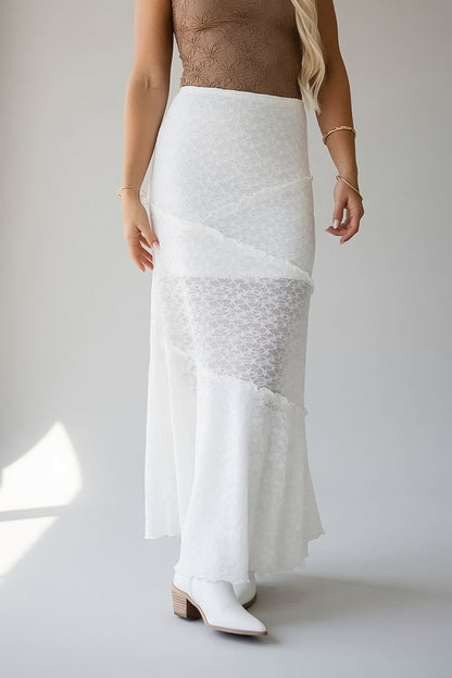 A woman modeling a white textured wrap-style maxi skirt with layered ruffle details and sheer patterned sections, paired with white pointed-toe boots against a light gray studio background.