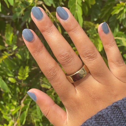 Hand with warm skin tone wearing Favorite Beanie slate grey gel polish manicure outdoors near green foliage background