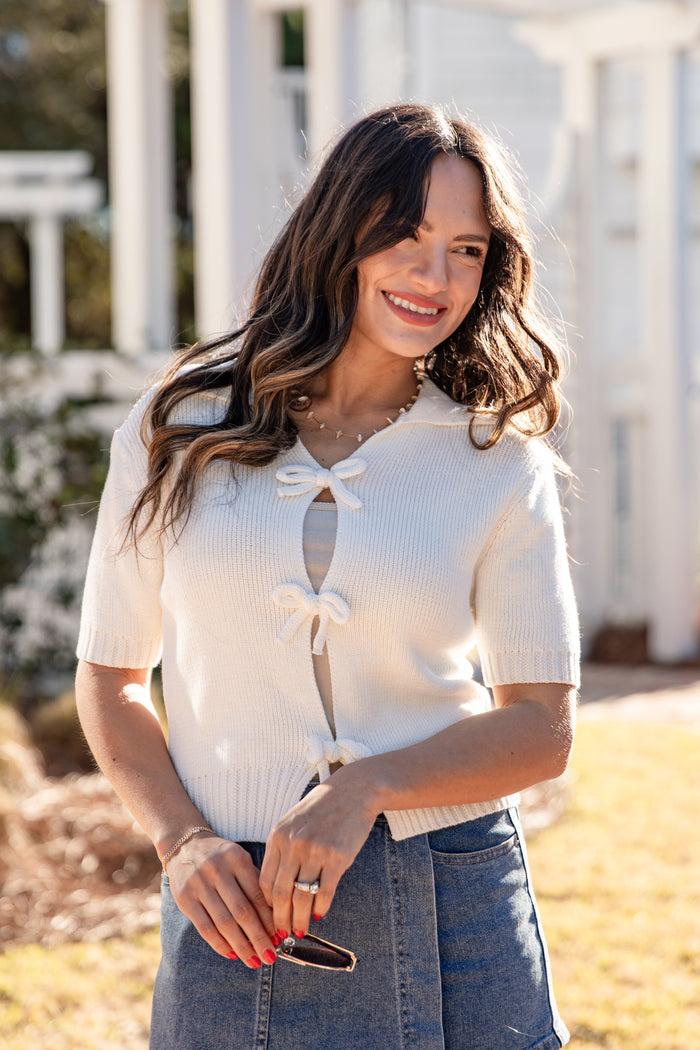 Woman wearing a white short-sleeve top with a bow detail, standing outdoors.