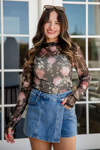 Woman wearing a floral long-sleeve top and denim skirt standing in front of a window.
