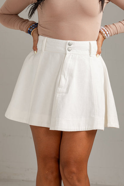 A model wearing a white high-waisted flared mini skirt with a buttoned waistband, paired with a beige long-sleeve top.