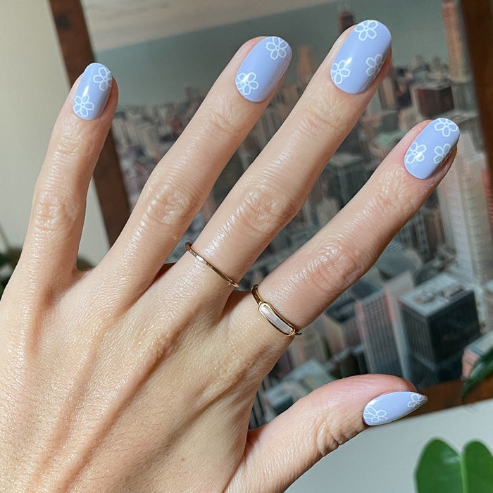 Floral Doodle | Short | Round - Tab Press-On nails in pastel blue with white flower doodles on hand with gold rings indoors