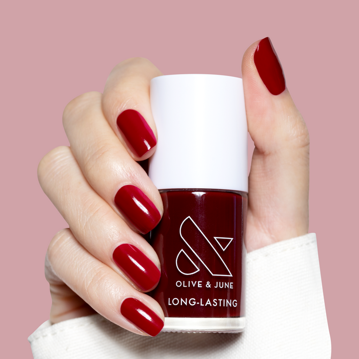Hand holding Garnet Glass nail polish bottle with deep scarlet gel polish on medium-length nails against pink background