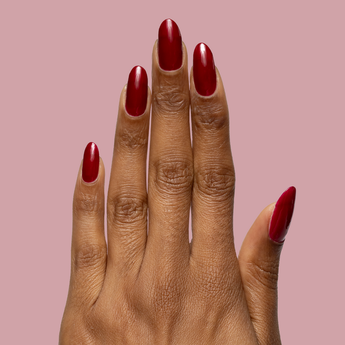 Hand with deep red glossy Garnet Glass gel polish on medium almond-shaped nails against muted pink background