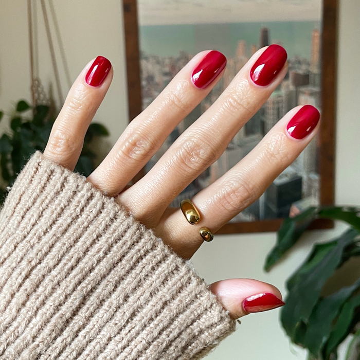Hand with medium-length nails painted in shiny Garnet Glass gel polish, wearing gold rings and a beige knit sleeve indoors