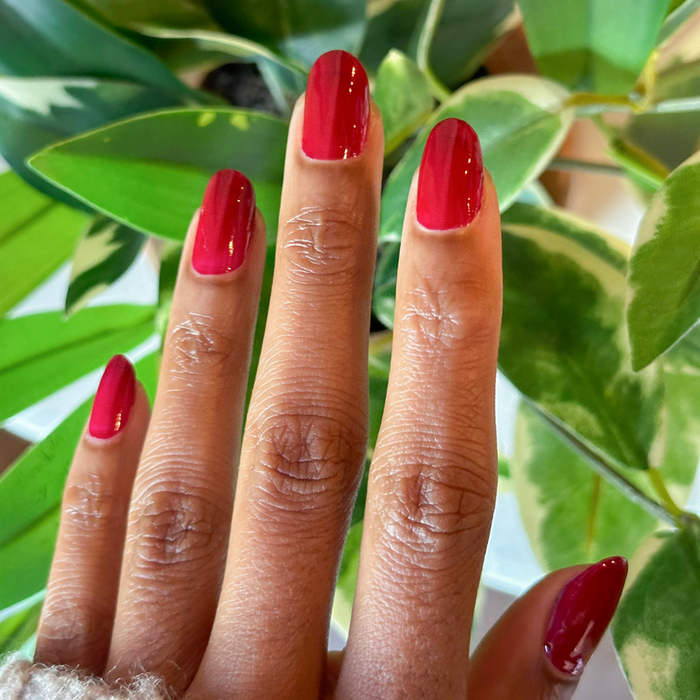 Fingers with medium-length nails painted in glossy Garnet Glass gel polish against green leafy background