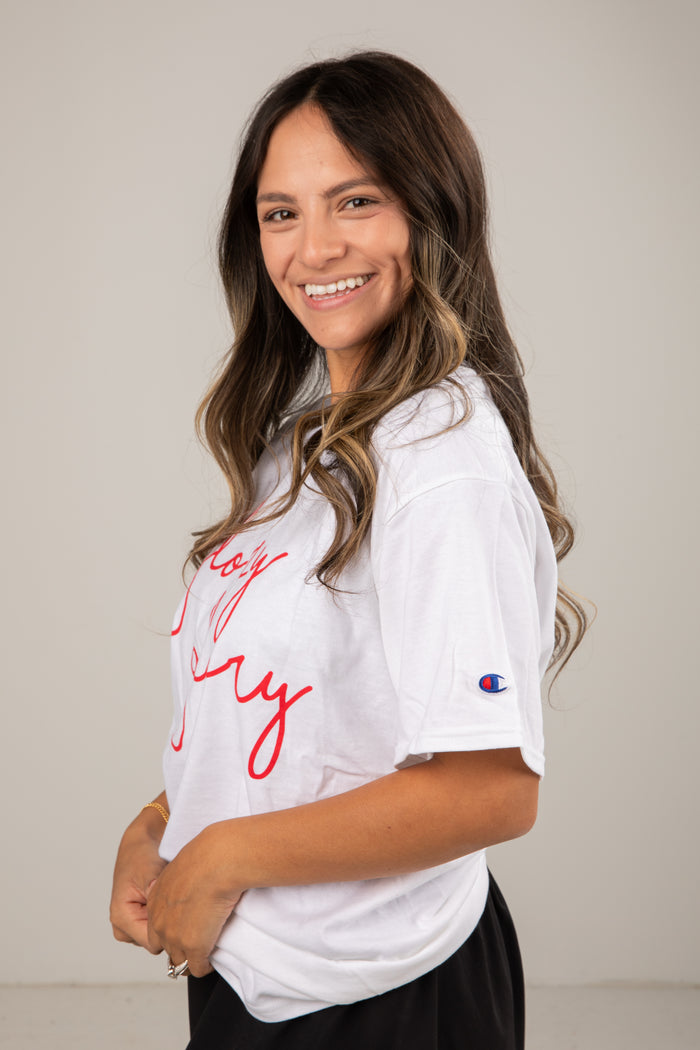 Woman wearing a white t-shirt with red text and a logo, standing against a plain background