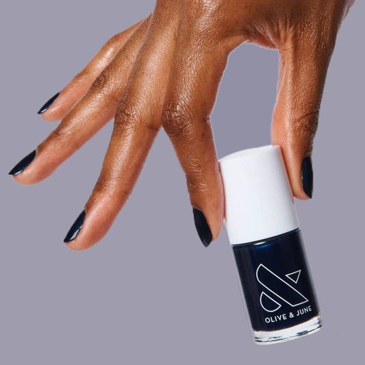 Hand with nails painted in shimmering inky navy holding HJ nail polish bottle by Olive and June for a polished mani