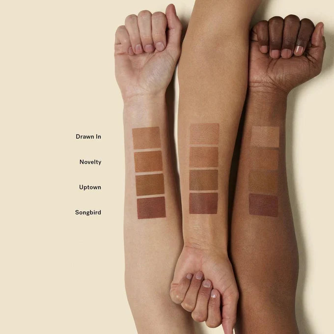 Swatch of ILIA Nightlite Bronzing Powder in Songbird showing warm, sun-kissed shades on arm, with product packaging visible.