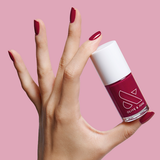 Hand holding a bottle of JG nail polish with nails painted in a rich berry shade against a pale pink background