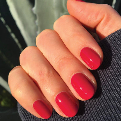 Close-up of hand showing smooth, glossy mani with JG polish in a bold berry shade against dark textured fabric background