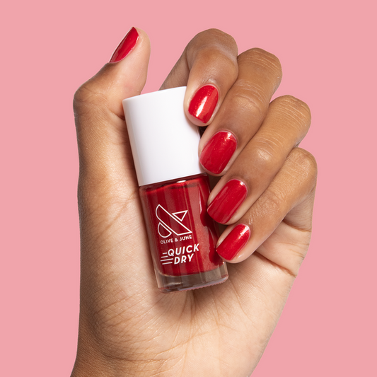 Hand with nails polished in glossy red shimmer Lippy polish holding Olive and June quick dry nail polish bottle against pink background
