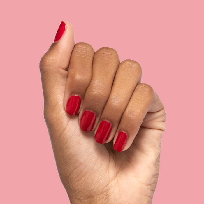 Hand with nails coated in glossy red shimmer Lippy polish showcasing a vibrant at-home manicure against light pink background