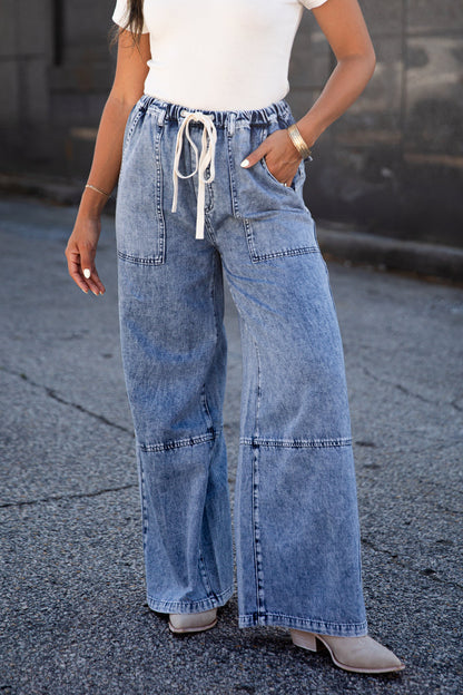 A person wearing light blue wide-leg denim jeans with a white drawstring waist, paired with a white top and beige heeled boots, standing outdoors on asphalt.