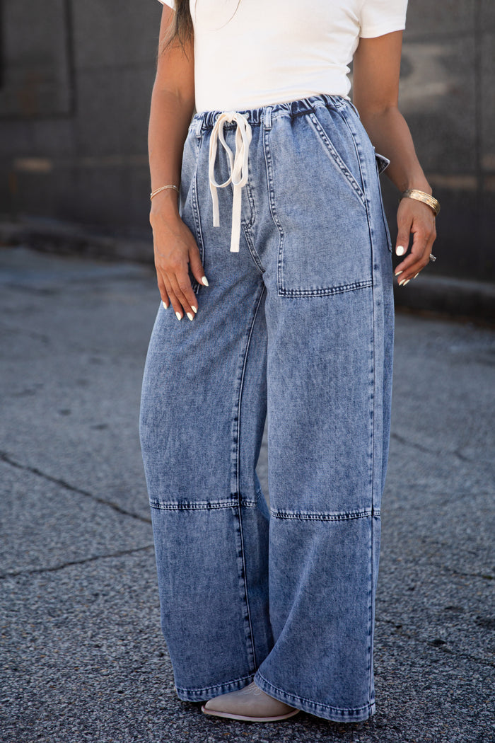 A person wearing wide-leg light blue denim jeans with a white drawstring waist, paired with a white top and gray shoes, standing outdoors on a paved surface.