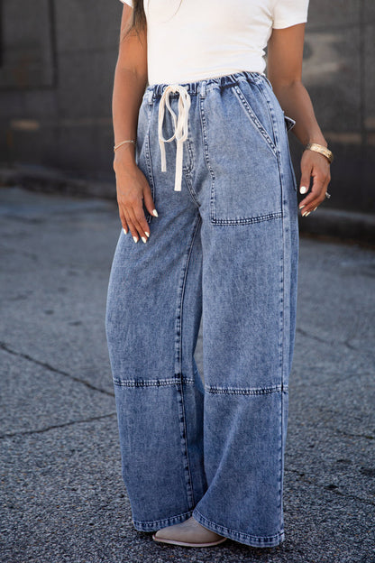 A person wearing wide-leg light blue denim jeans with a white drawstring waist, paired with a white top and gray shoes, standing outdoors on a paved surface.