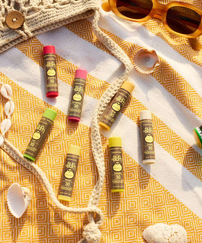 Original SPF 30 Sunscreen Lip Balm - Pineapple displayed on a yellow patterned beach blanket with seashells and yellow sunglasses, featuring colorful lip balm tubes arranged in a casual beachy layout - by Sun Bum
