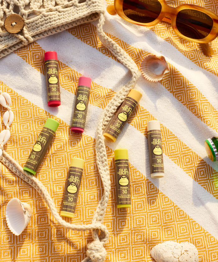 Original SPF 30 Sunscreen Lip Balm in Watermelon flavor displayed on a yellow patterned beach blanket with seashells and sunglasses, showcasing multiple colorful lip balm tubes in a summer beach setting - by Sun Bum