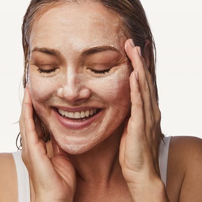 A close-up of a woman with a white clay or face mask applied, smiling with her eyes closed and her hands resting on her cheeks against a clean white background.