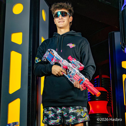 Person wearing a black hoodie and colorful shorts holding a Nerf gun in front of a colorful background.