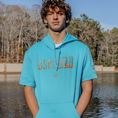 Person wearing a turquoise hoodie with 'BSBLSZN' text standing by a body of water with trees in the background