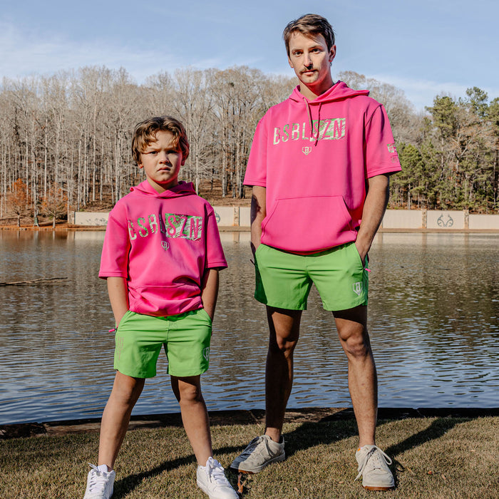 Two people wearing matching pink hoodies with 'BSBL SZN' and green shorts standing by a body of water.