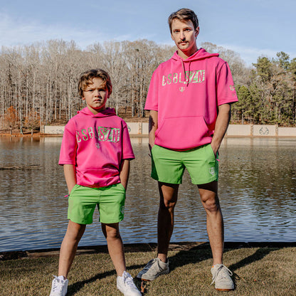 Two people wearing matching pink hoodies with 'BSBL SZN' and green shorts standing by a body of water.