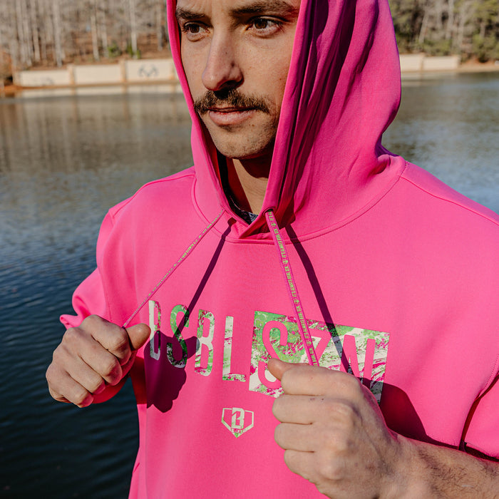 Person wearing a bright pink hoodie with 'BSBL SZN' text, standing by a body of water.