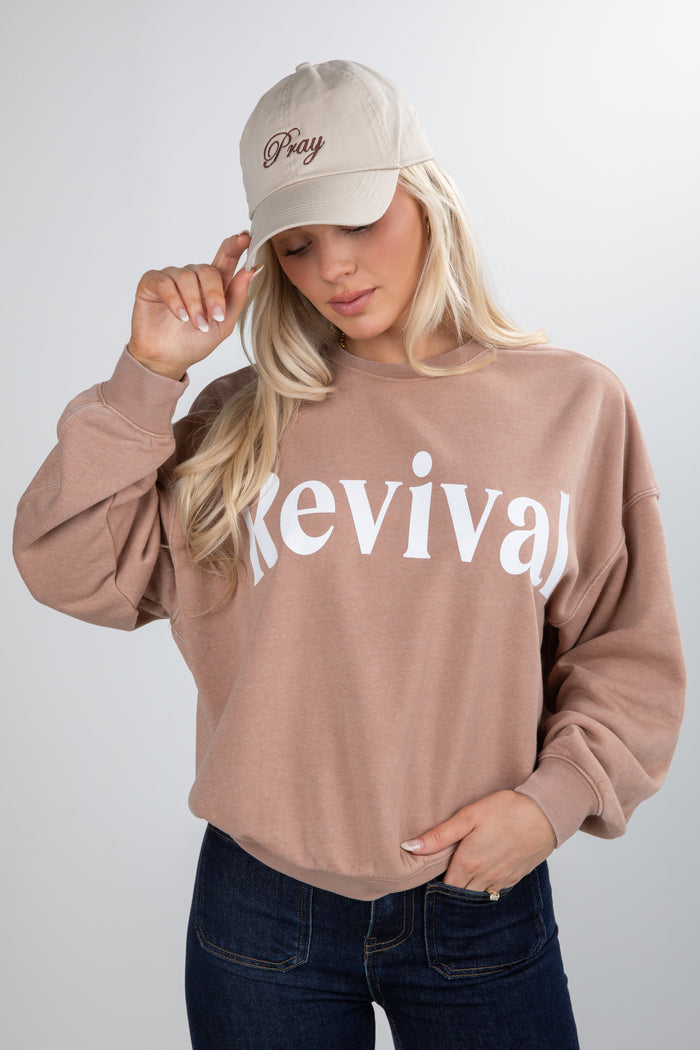 Person wearing a beige sweatshirt with 'Revival' text and a matching cap on a plain background
