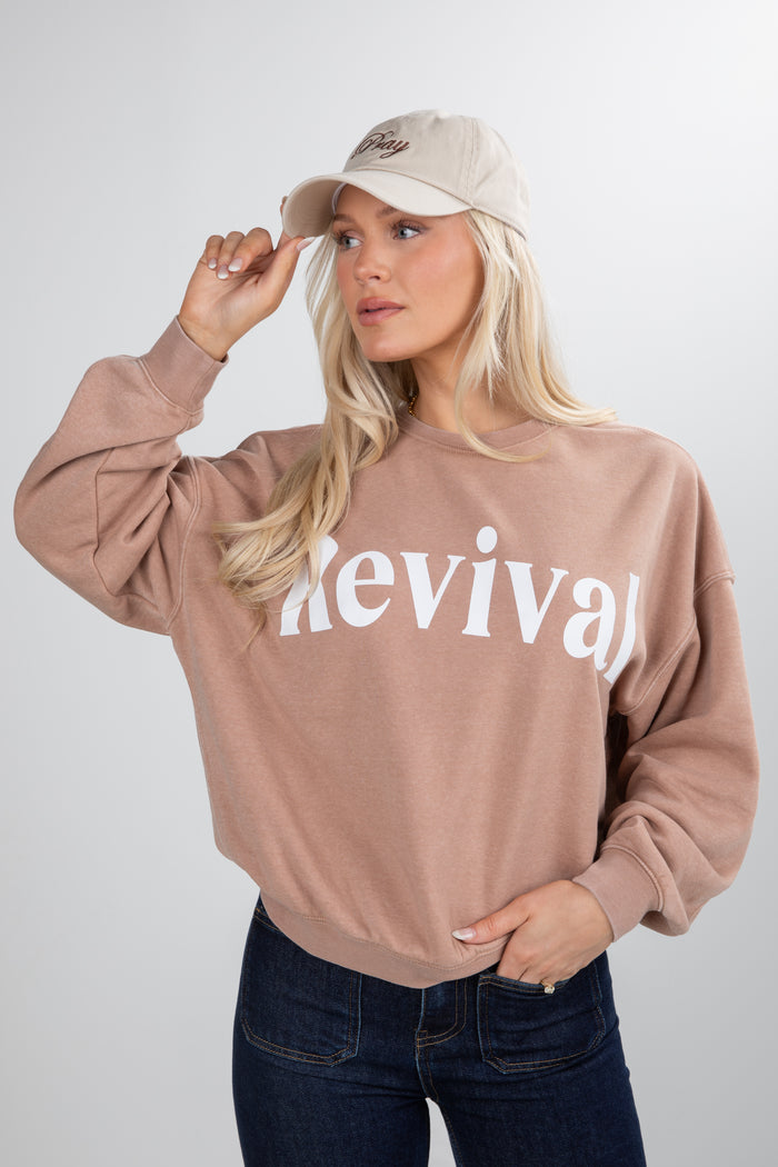 Woman wearing a beige sweatshirt with 'Revival' text, holding a beige cap.