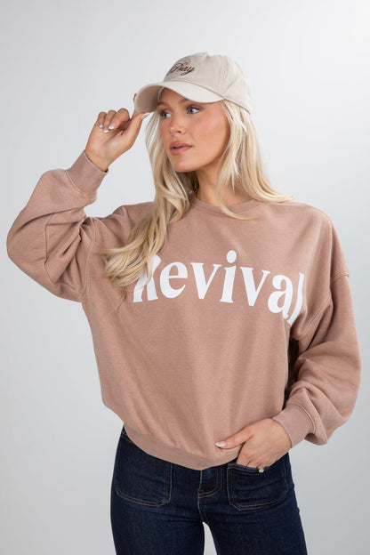 Woman wearing a beige sweatshirt with 'Revival' text, holding a beige cap.