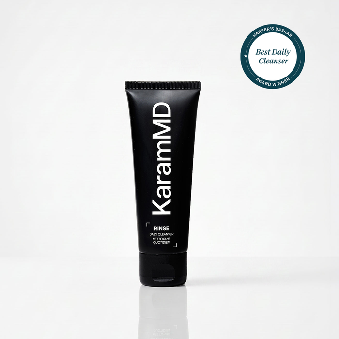 A black squeeze tube of KaramMD Rinse Daily Cleanser standing upright on a light gray reflective surface. A circular teal award badge reading Harper's Bazaar Best Daily Cleanser Award Winner appears in the top right corner.