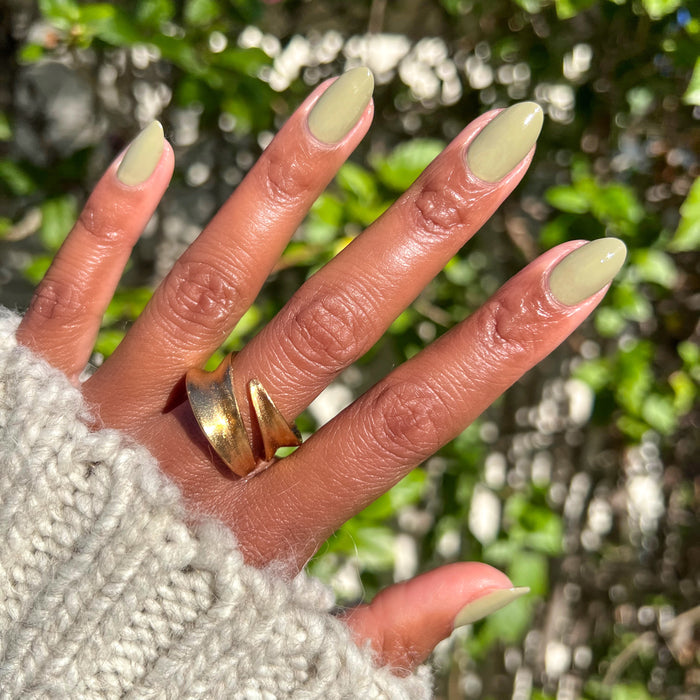 Hand with nails painted in soft Sagey polish showing a manicure with warm natural lighting and gold rings