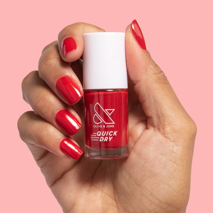 Hand holding a bottle of Smooch quick dry nail polish with matching red painted nails on pink background