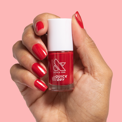 Hand holding a bottle of Smooch quick dry nail polish with matching red painted nails on pink background