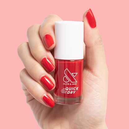 Hand holding a bottle of Smooch nail polish with nails painted in rich scarlet red quick dry polish on pink background