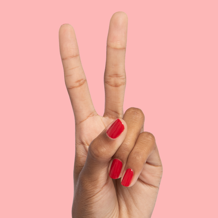 Hand with nails painted in glossy red Smooch polish showing a peace sign on a soft pink background
