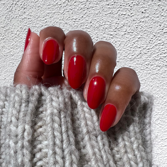 Hand with long nails painted in glossy rich scarlet red Smooch polish against textured white background and cozy knit sleeve